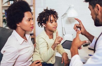 A young girl communicates to a male doctor using sign language while her mother looks on. They are in the doctor's office. The girl is wearing a yellow top.