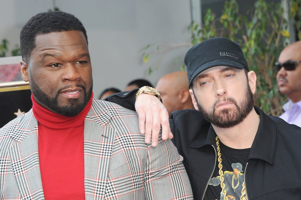 50 Cent in a plaid suit and Eminem in a casual outfit with a chain necklace pose together, with Eminem's arm on 50 Cent's shoulder