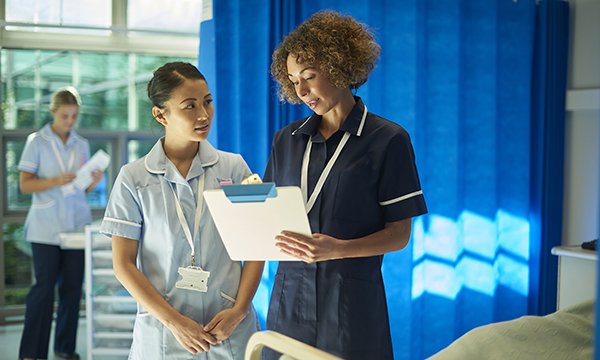 A band 7 nurse on a hospital ward checks a patient’s notes and guides a junior nurse team member