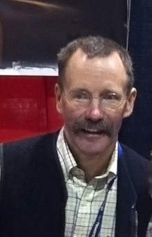 A middle-aged caucasoid man looking at the photographer; he is presented as a bust, wearing a yellow-plaid collared shirt under a cardigan sweater.