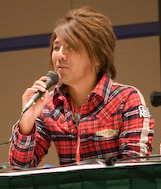 Portrait d'un homme brun d'origine japonaise habillé d'une chemise rouge et noire à carreaux, avec un micro dans la main qu'il porte à sa bouche.
