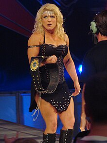 Beth Phoenix portant une tenue de ring et une ceinture de championne dans la main droite.