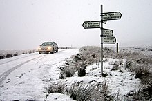Photographie couleur de Sally Gap sous la neige avec une voiture, phares allumés, passant au niveau d'un panneau indiquant plusieurs directions et distances