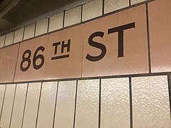 Detailaufnahme. 86th Street, Stationsname in Dunkelbraun auf ockerfarbenem Farbstreifen, senkrechte helle Fliesen mit dunkelgrauen Fugen