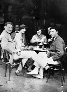 three men, dressed in light colored trousers and wearing hats, and two women, wearing light colored dresses, sitting at a sidewalk table