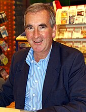 A photograph of a man in his fifties looking at the camera. He is wearing an open-collared blue shirt and a blue jacket.