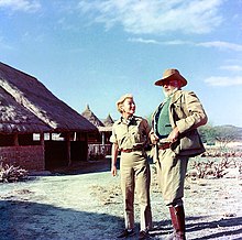 photograph of a man and woman with a brush covered hut in the background