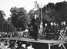 "Black and white photograph of Michael Collins in a suit standing on a makeshift stage with people sitting behind and in front of him"