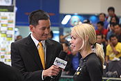 a male reporter in a suit with a microphone, speaking with Ouschan on the tournament floor