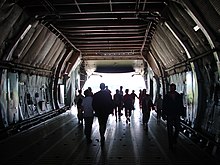 Plusieurs personnes en contre-jour debout dans la soute, la rampe arrière de l'avion est ouverte.