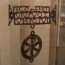 A metal plate with holes. The outline spells 'EGOZENO / VIVSVOT / VM POSVI'. A Chi-Rho shape hangs from it.