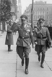 Photographie en noir et blanc d'un homme en uniforme, saluant, suivi dans la rue par un autre officier.