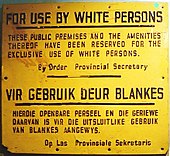 A yellow sign with black letters that states, in English: "FOR USE BY WHITE PERSONS. THESE PUBLIC PREMISES AND THE AMENITIES THEREOF HAVE BEEN RESERVED FOR THE EXCLUSIVE USE OF WHITE PERSONS. By Order Provincial Secretary"
