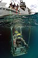 Two divers wearing lightweight demand helmets stand back-to-back on an underwater platform holding on to the railings. The photo also shows the support vessel above the surface in the background. (from Outline of underwater diving)