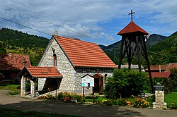 A picture of a small white stone church with an orange tile roof. The sun is high overhead and flowing plants are growing just in front of the church. A second building and green hills are visible in the background. A cross is above the church.