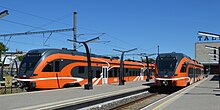 Seitliche Farbfotografie von einem Bahnsteig mit zwei roten Zügen. Auf den Zügen verlaufen weiße Linien. Im rechten Hintergrund sind auf einem Dach die ersten drei Buchstaben von Tallinn zu sehen.