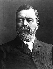 A black and white portrait photo of a thickly bearded man in a wool coat.