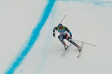 Photographie d'un skieur dans la descente, jambes fléchies, portant une combinaison grise et noire et le dossard numéro 16.