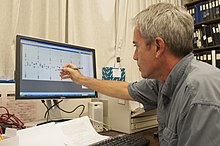 Bernard Crespi (the primary originator of the imprinted brain hypothesis), a man with short grey hair in a grey shirt seen from behind, points at a computer screen with a pen. The screen appears to be showing an image of a genome being analyzed.