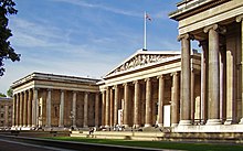 Seitliche Farbfotografie eines braunen Gebäudes mit Seitenflügeln, die durchgehend aus Säulenvorhallen bestehen. Der Mittelteil hat ein Giebelfeld mit antiker Szene und einer britischen Flagge auf dem Dach. Auf den vorderen Stufen sitzen Menschen.