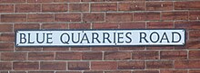 On a red brick wall, a rectangular sign bears the name "Blue Quarries Road" in black, upper case letters on a white background.