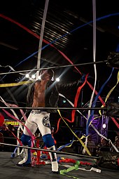 Catcheur en pantalon blancsaluant la foule les bras écartés et recevant des rubans de couleurs pour son accueil sur le ring.