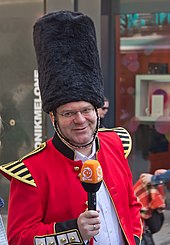 Abgebildet ist der Moderator Elton, gekleidet in eine an französische Uniformen des späten 19. Jahrhunderts erinnernde Karnevalsuniform des Kölner Karnevals it einem hohen Fellhut