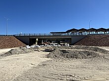 A road elevated on an embankment with a pedestrian underpass under construction at ground level. The station's shelter can be seen behind the road in the background.