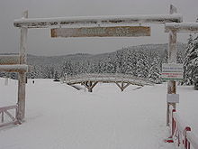 Clairière enneigée avec au premier plan un portail en bois et un insigne « Autrans : bienvenue à Gève, accès aux pistes », au second plan un ponton en bois et tout autour des conifères lourdement enneigés