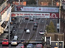 High level shot of multi lane road lower down stretching into distance with two glass sided pedestrian walkways over linking car park to right and shopping centre to left