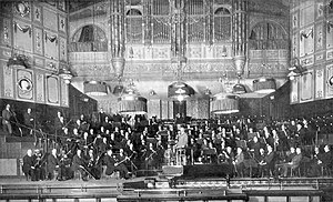 large orchestra and their conductor seen on the platform of Victorian concert hall in long shot
