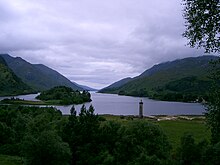 A gloomy, cloud-covered sky above a large lake surrounded by green hills. There is a wooded islet at left and a tall circular tower in the foreground topped by a statue.
