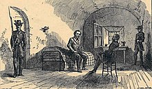 Dessin montrant un homme en costume assis sur un lit rustique dans une salle dépouillée au plafond voûté. Il discute avec un homme assis sur une chaise devant lui tandis que deux soldats portant un fusil à baïonnette montent la garde dans la cellule.