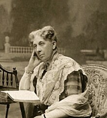 A sepia photograph showing the head and shoulders of a seated woman beside a small table on which is an open book