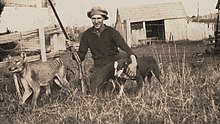 Černobílá fotografie farmáře, jenž pózuje se psem, puškou a vycpaným vakovlkem