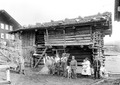 Photographie noir/blanc où posent plusieurs soldats et paysans, une paysanne portant son enfant, devant une grange ; deux paysans portent des fagots de foin et un autre une hotte