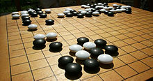 Linsenförmige Spielsteine in Schwarz und Weiß liegen unregelmäßig auf einem Holzbrett verteilt, das horizontal und vertikal liniert ist.