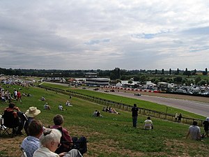 View of the track stretching away to the left from an elevated grassy bank with spectators, showing circuit buildings of far side of track with lakes beyond against a dappled sky
