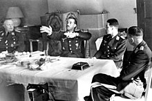 Black-and-white photograph of four men wearing uniforms sitting on wooden chairs around a table in a living room. An older man is sitting at the head of the table on the left. Two younger men are sitting along the table's side, with their backs toward a tiled fireplace on the room's far wall. A fourth man is sitting at the head of the table on the right, leaning back, his left leg folded over his right. The table is covered by a white table cloth. The right-hand side of the table is empty, except for a large dark ashtray, a spoon i an empty glass; the left i centre of the table is covered by an assortment of empty plates, coffee cups i other dishes; there is also a bowl of fruit. The second man from the left has his hands raised, palms facing the camera, i the fingers of both hands pointing to the left of the image. The heads of the three other men are turned toward him.