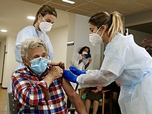 Elderly woman rolls up sleeve as two nurses administer a vaccine