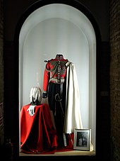 Photographie en couleur montrant un uniforme rouge et noir exposé sur un mannequin sans tête, dans une vitrine en forme d'arche.
