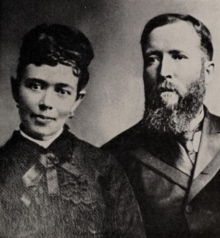 A portrait photograph of Ida Frances Hunt Udall (on the left) and David King Udall (on the right). Ida's hair is done up. She wears earrings, and the dress she wears features an ornate bow at the collar. David wears a necktie and suit, which is buttoned, and he has a long beard that reaches past his collar.