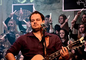 Ondřej Ládek wearing a burgundy shirt, playing an acoustic guitar in front of a microphone, with young girls holding candles or glow sticks in the background