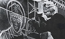 Edward Catich, a white man with glasses and light hair, uses a hammer and chisel to carve an image of a religious figure into a wall