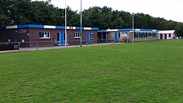 Op de bovenste foto is het entree van het sportpark te zien. Op de onderste foto zijn de kleedkamer (links) en de kantine (rechts) te zien.