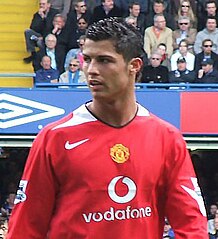 Head and torso of a man wearing a red long-sleeved football shirt. He is in front of a crowded stand of fans.