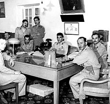 Huit hommes en uniformes militaires posent dans une pièce autour d'une table rectangulaire. Tous sauf deux sont assis.