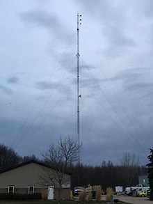 An alternating white and red triangular guyed lattice tower, set against a cloudy sky. A three-bayed antenna at the top of the structure is also flanked by an activated red beacon light. An additional red beacon light is fixed on the structure above a small studio-to-transmitter link dish. An office building is pictured in front of the tower on the left-hand side, with a parking lot on the other side.