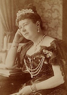 A sepia-toned, formal photograph of a seated, bejewelled woman