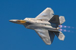 A pilot peers up from his F-22 Raptor while in-flight, showing the top view of the aircraft. The terrain of Nevada can be seen below mostly cloudless skies. Aircraft is mostly gray, apart from the dark cockpit canopy.